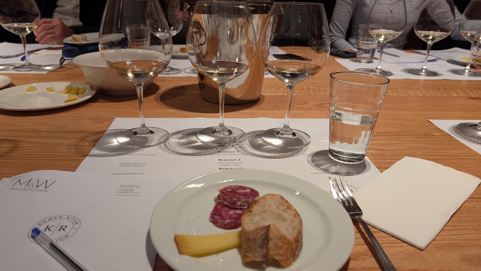 A place setting at the long table, the first bracket poured into three glasses standing on a printed placemat, beside a plate of cheese, salami and bread
