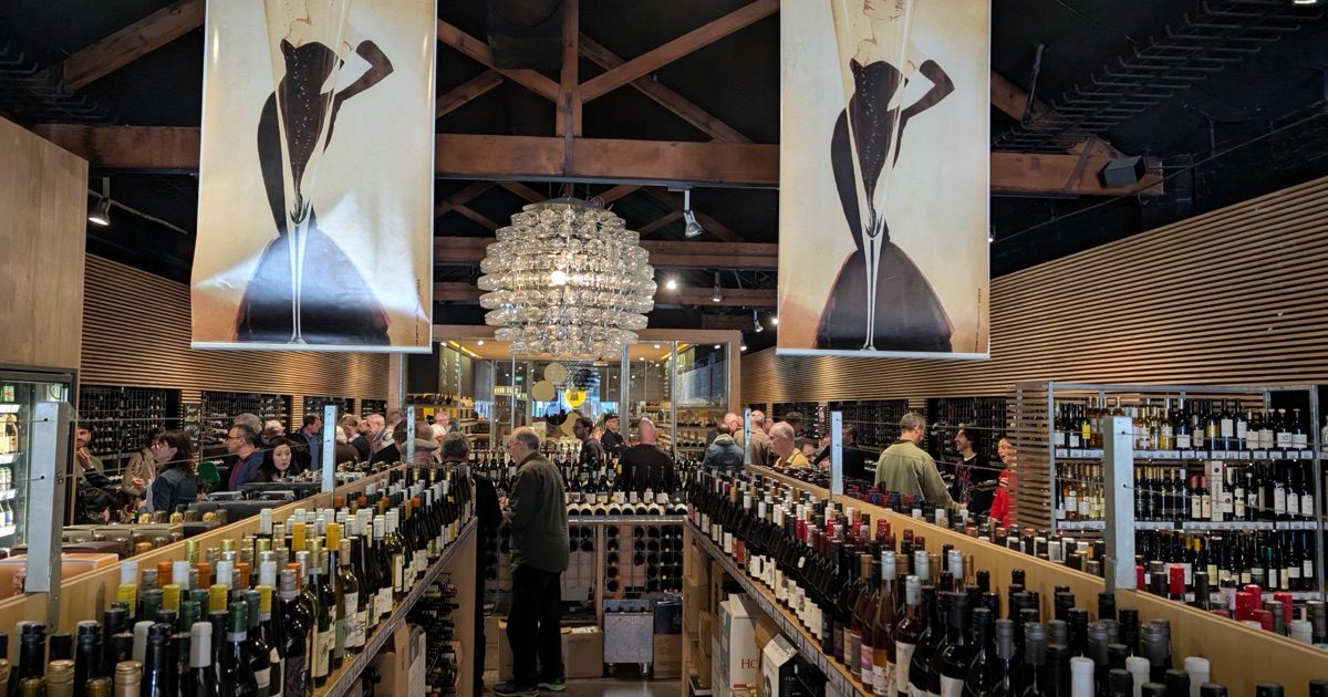 The main room at Prince Wine Store during the tasting, rows of bottles in the foreground and a crowd gathered at the far end beneath a chandelier made of wine glasses