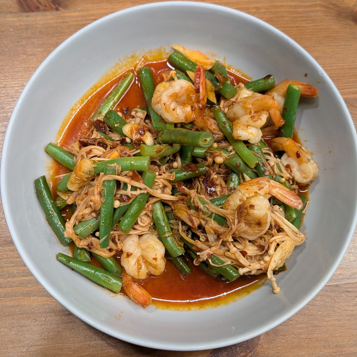 A bowl of prawns stir-fried with green beans and mushrooms in a red chilli sauce, on a wooden table