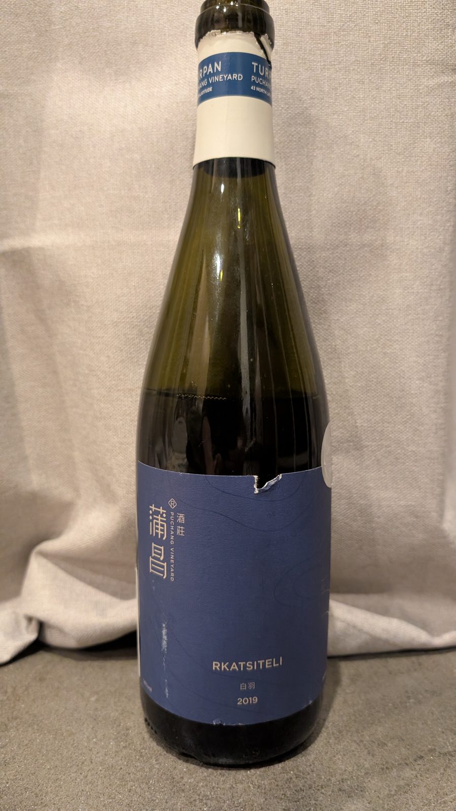 A bottle of Puchang Vineyard Rkatsiteli 2019 with a blue label and white capsule, standing against a cream curtain