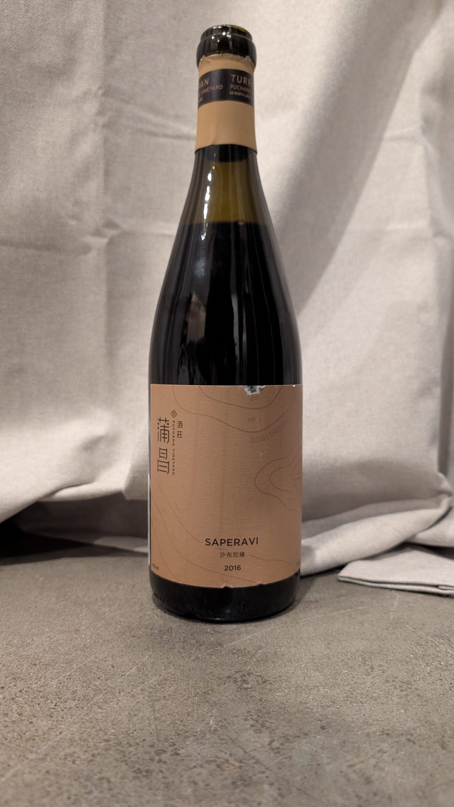 A bottle of Puchang Vineyard Saperavi 2016 with a sand-coloured label and capsule, standing against a cream curtain