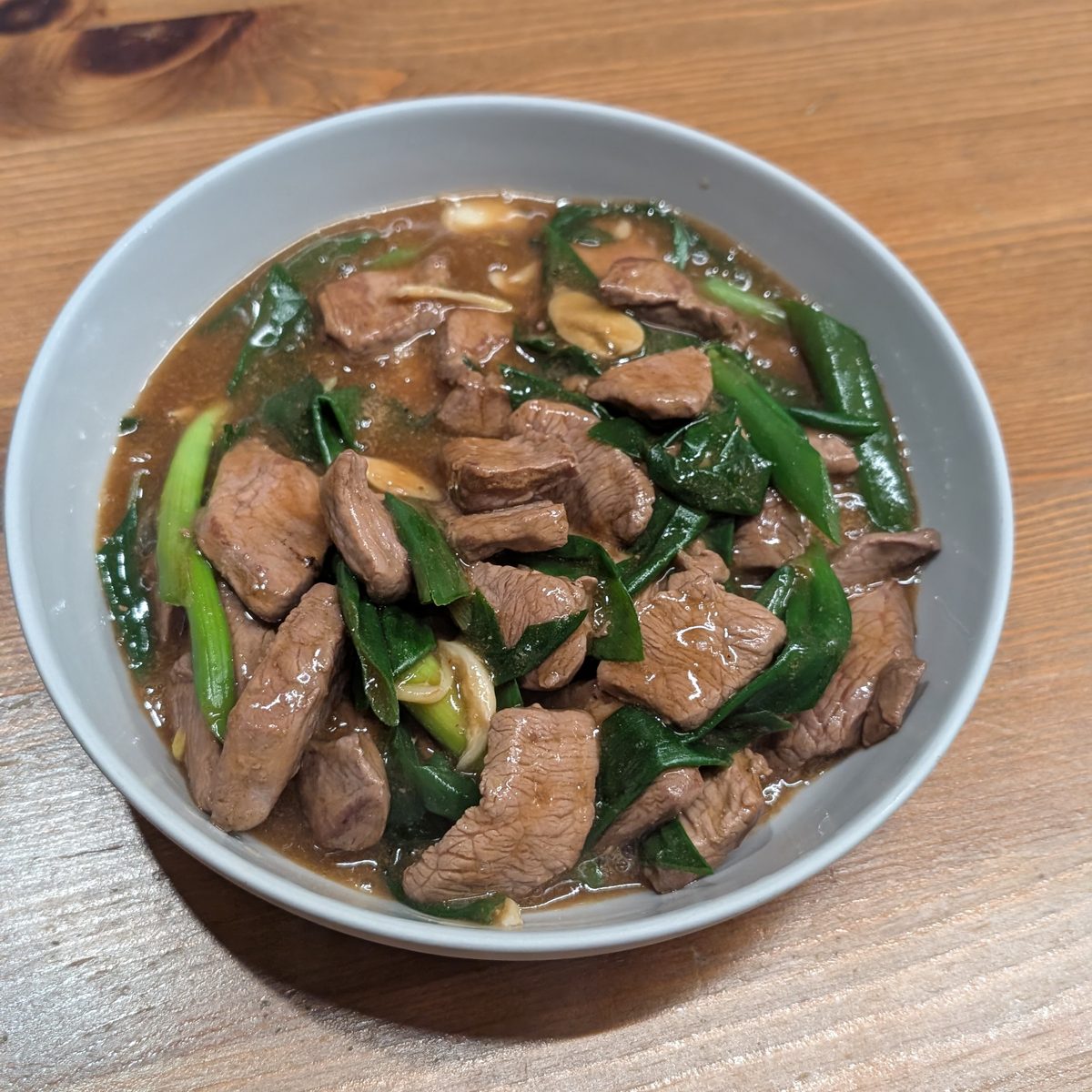 A bowl of sliced venison stir-fried with spring onions and leafy greens in a dark sauce, on a wooden table
