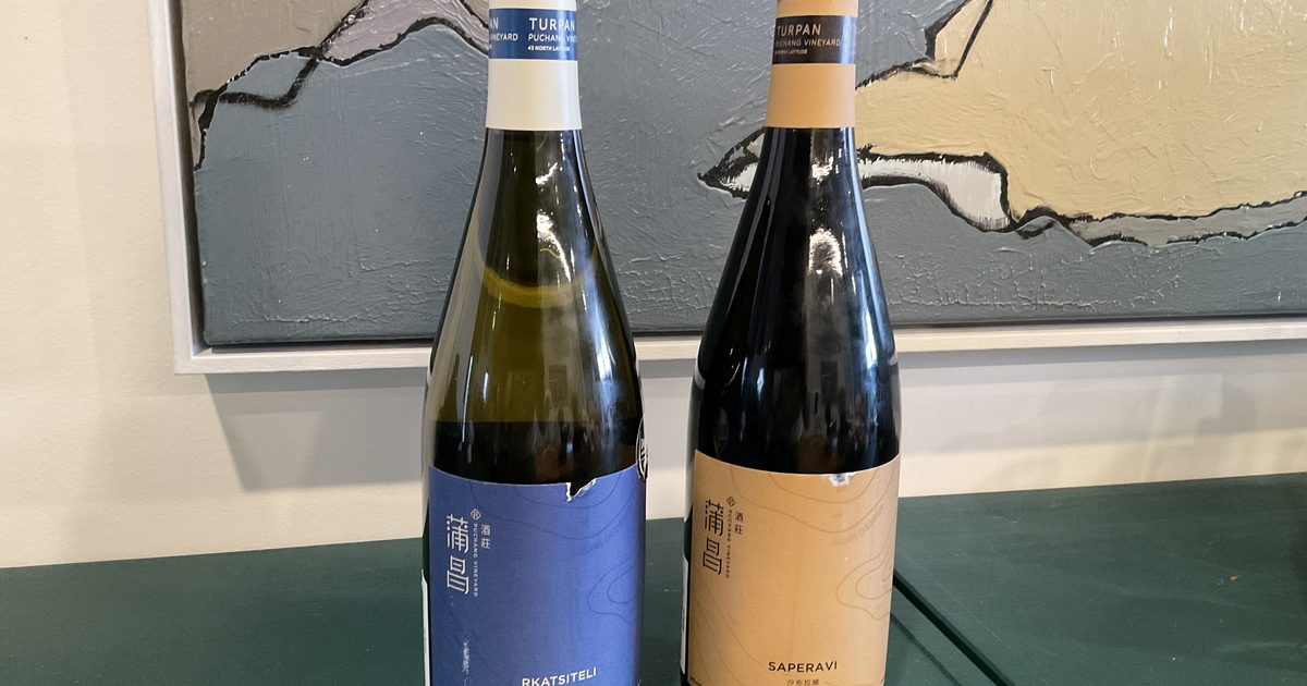 Two Puchang Vineyard bottles on a green table in front of a painting, a white Rkatsiteli with a blue label and a red Saperavi with a sand-coloured label, both with neck labels reading Turpan