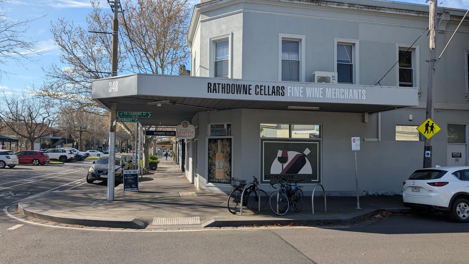 The Rathdowne Cellars shopfront on a street corner in Carlton North under a clear blue sky, its awning signed Rathdowne Cellars Fine Wine Merchants, with bicycles parked outside