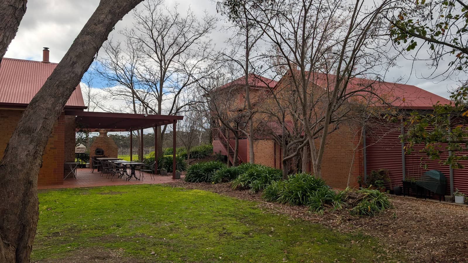 The winery buildings in red brick under red corrugated roofs, a covered terrace with tables and a wood-fired oven on the left and bare winter trees between them