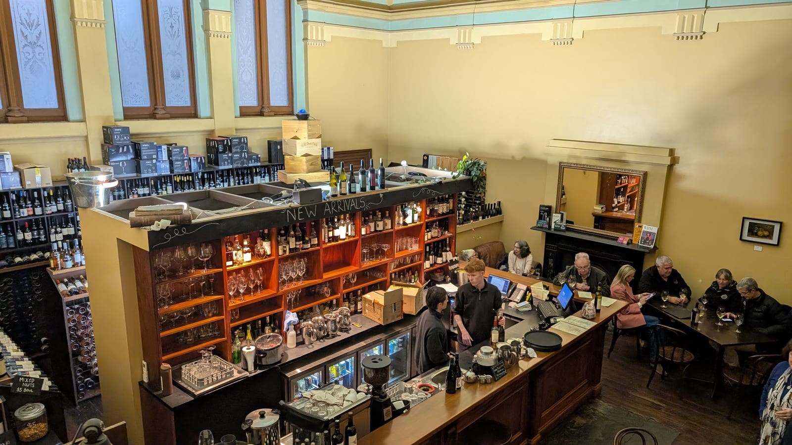 The bar and shop floor at Wine Bank on View seen from the mezzanine, a long timber counter backed by lit shelves of glassware and spirits, under the high ceiling and arched windows of the old banking chamber